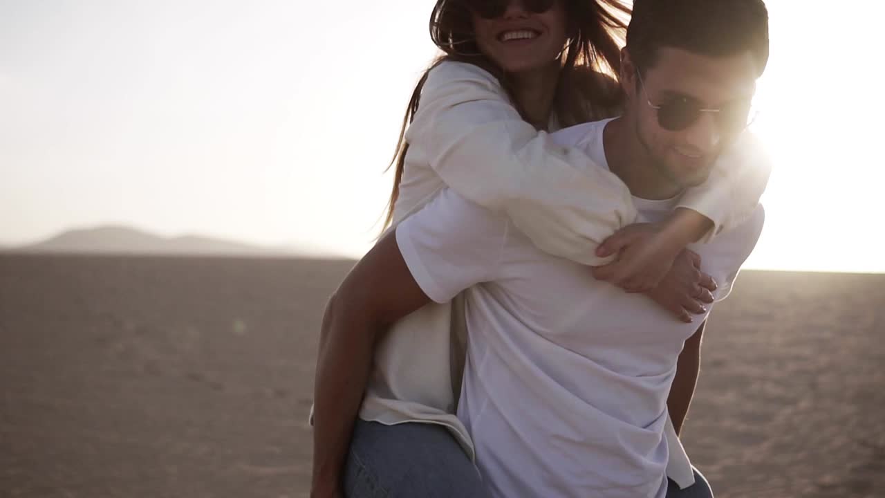 feliz y linda adorable pareja adulta en camisas blancas y vaqueros hombre con mujer novia en la espalda de cerdo, divertirse jugar, reír, sonreír en la puesta de sol en el desierto loco en el amor, las emociones y la relación