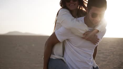 happy and cute adorable adult couple in white shirts and jeans man with woman girlfriend on piggy back, have fun play, laugh,smile on sunset at desert crazy in love, emotions and relationship