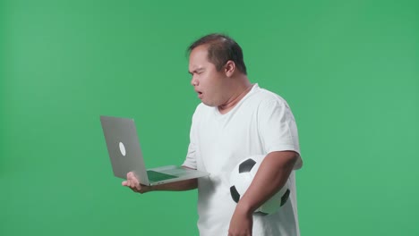 vista lateral de un hombre asiático con una pelota se arrepiente de animar el fútbol en una computadora portátil en el estudio de pantalla verde