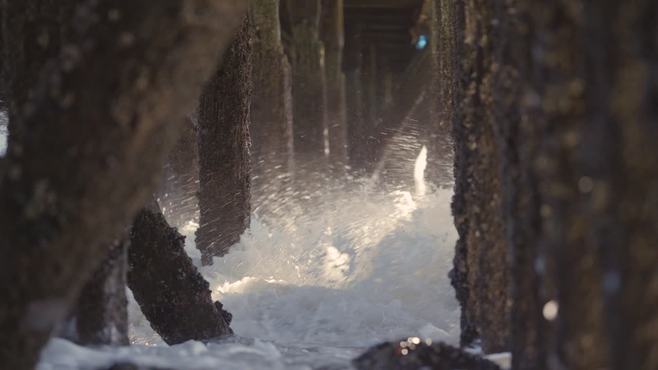 Waves crashing under a pier, old wooden pillings with sunbeams coming through