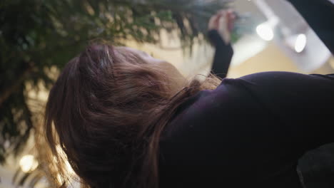 woman with curly brown hair in black outfit stretches arm upward to gently touch green leaf while looking up under indoor tree, framed by moody lighting and soft natural shadows from plant leaves