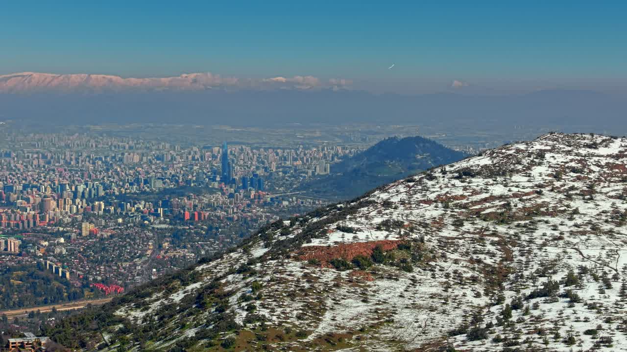 Establishing aerial view of the snow-capped peak of Mount Manquehue and the developed city of Santiago, Chile, with two airplanes flying over the city on a clear, sunny day