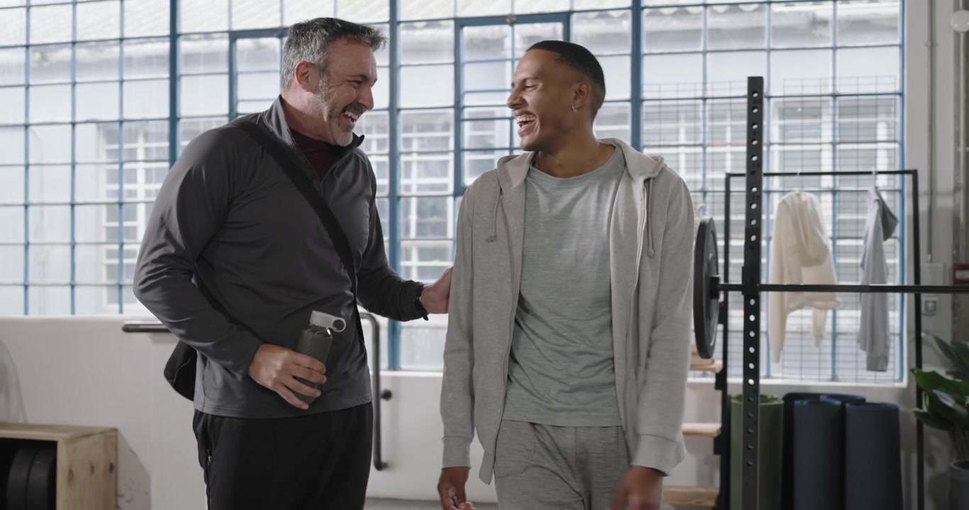 Diverse male friends greeting and laughing in gym as man with water bottle approaching barbell