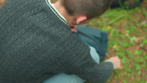 rear view of man adjusting tent bag and zipping it shut on grassy woodland floor while another bag lies nearby, suggesting camping activity in quiet outdoor natural forest environment