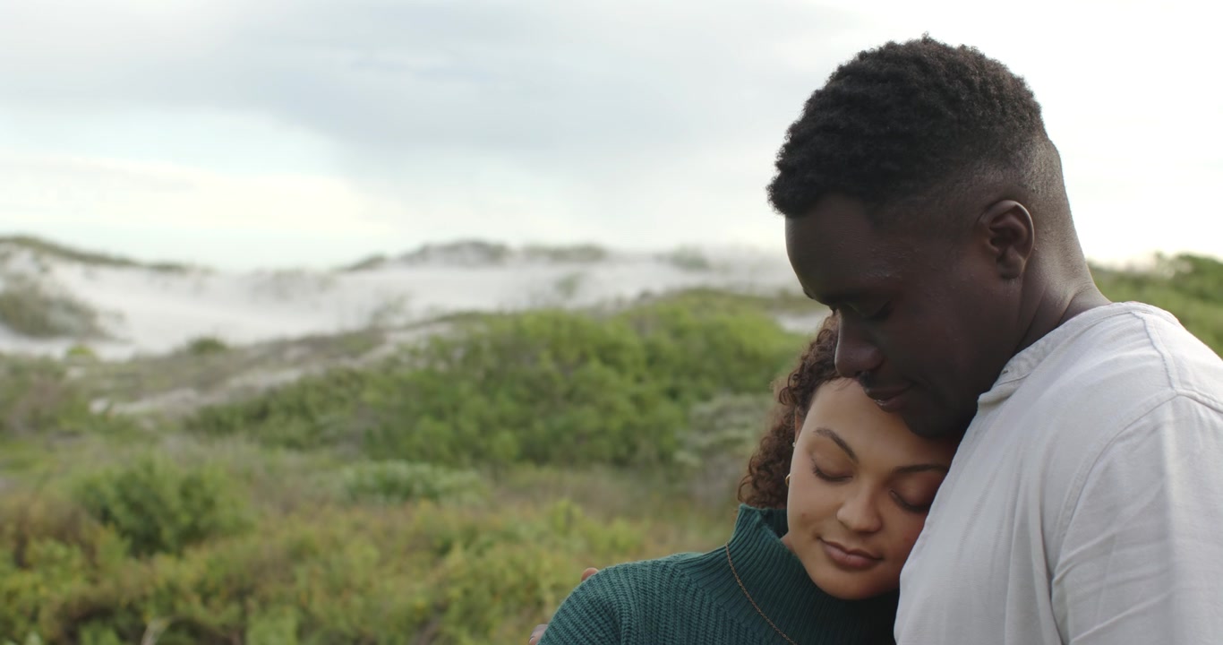 Coppia afroamericana che si abbraccia e si conforta sulle dune, con la telecamera che zooma su un top leggero in maglia color verde acqua, con spazio per il testo.