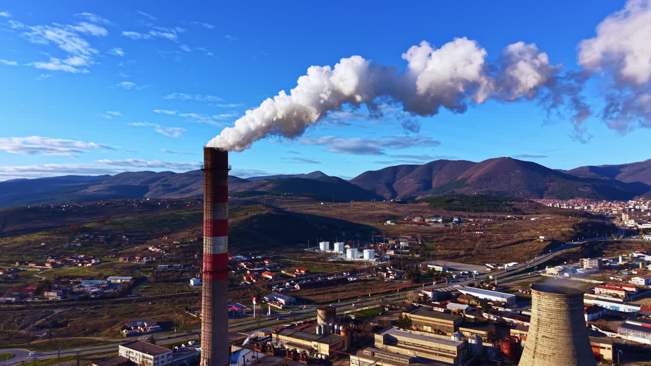 Il fumo si innalza da una ciminiera di fabbrica in una regione montuosa durante le ore diurne, mostrando l'attività industriale e il paesaggio circostante.