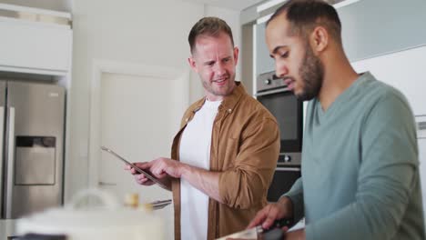 Multi-ethnic-gay-male-couple-preparing-food-in-kitchen-one-using-tablet