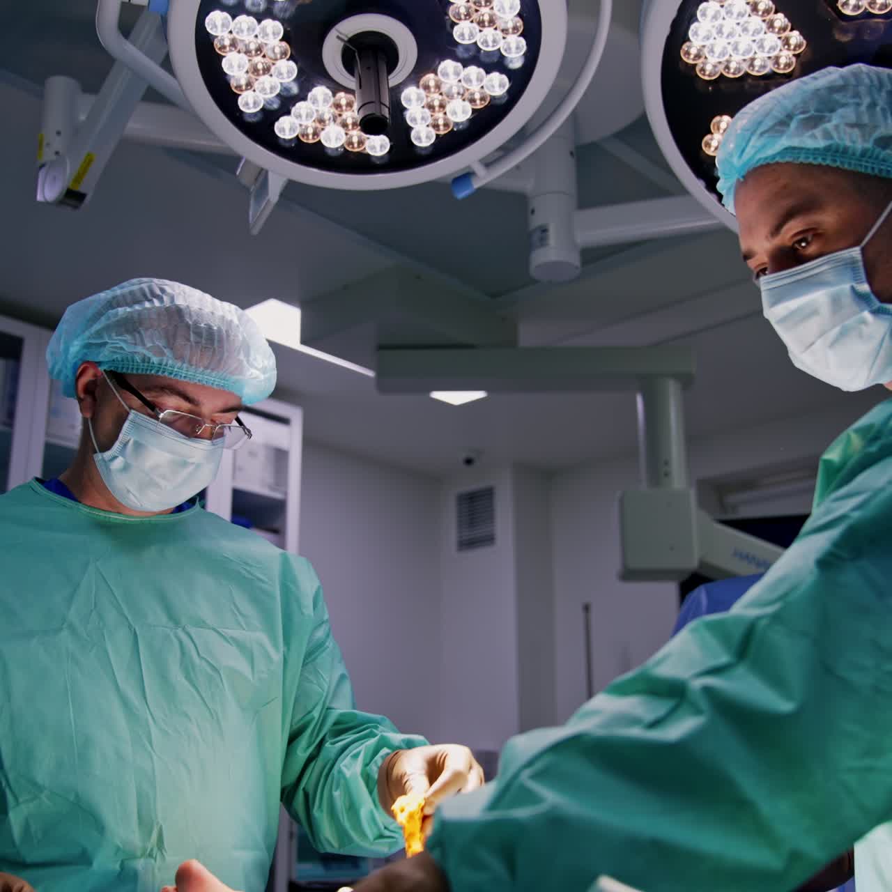 Læger opererer en patients traumatiserede fod. Runde lamper er tændt bag kirurgerne. Lav vinkel.