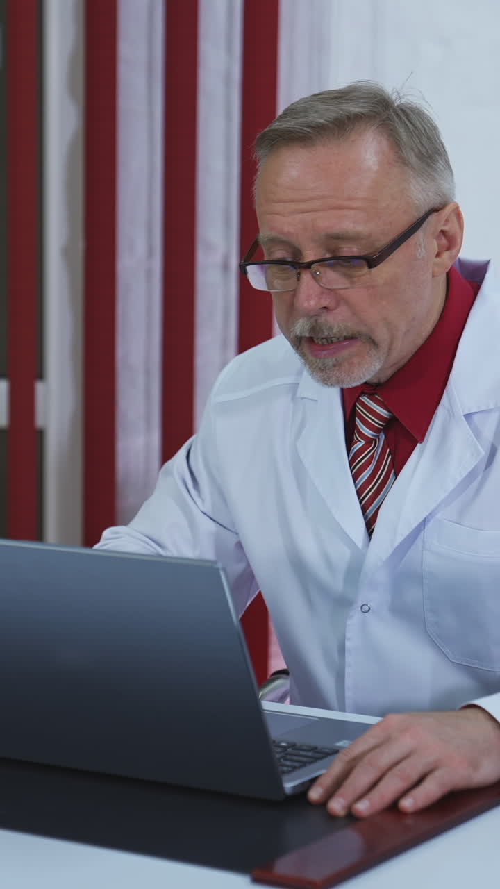 Older doctor making video call on laptop. Senior male physician talking to patient while looking at pc screen communicating by webcam in web chat. Telemedicine concept. Vertical video