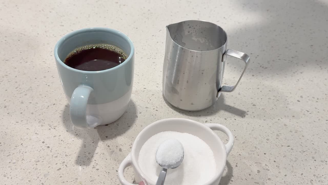 A hand adds sugar to a coffee mug on a kitchen counter, with a milk jug nearby