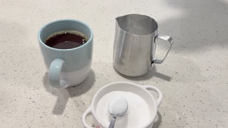 a hand adds sugar to a coffee mug on a kitchen counter, with a milk jug nearby