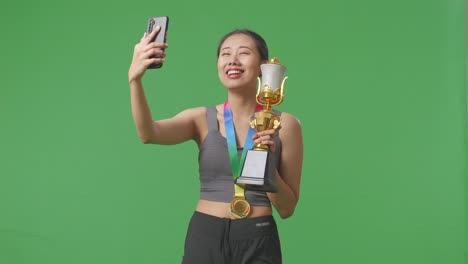 asian woman with a gold medal showing a gold trophy to the video call on smartphone on green screen background in the studio