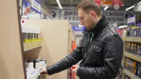 young man choosing spray paint in dye shop. rows of shelves with paint cans. he is shaking a canister and looking at label