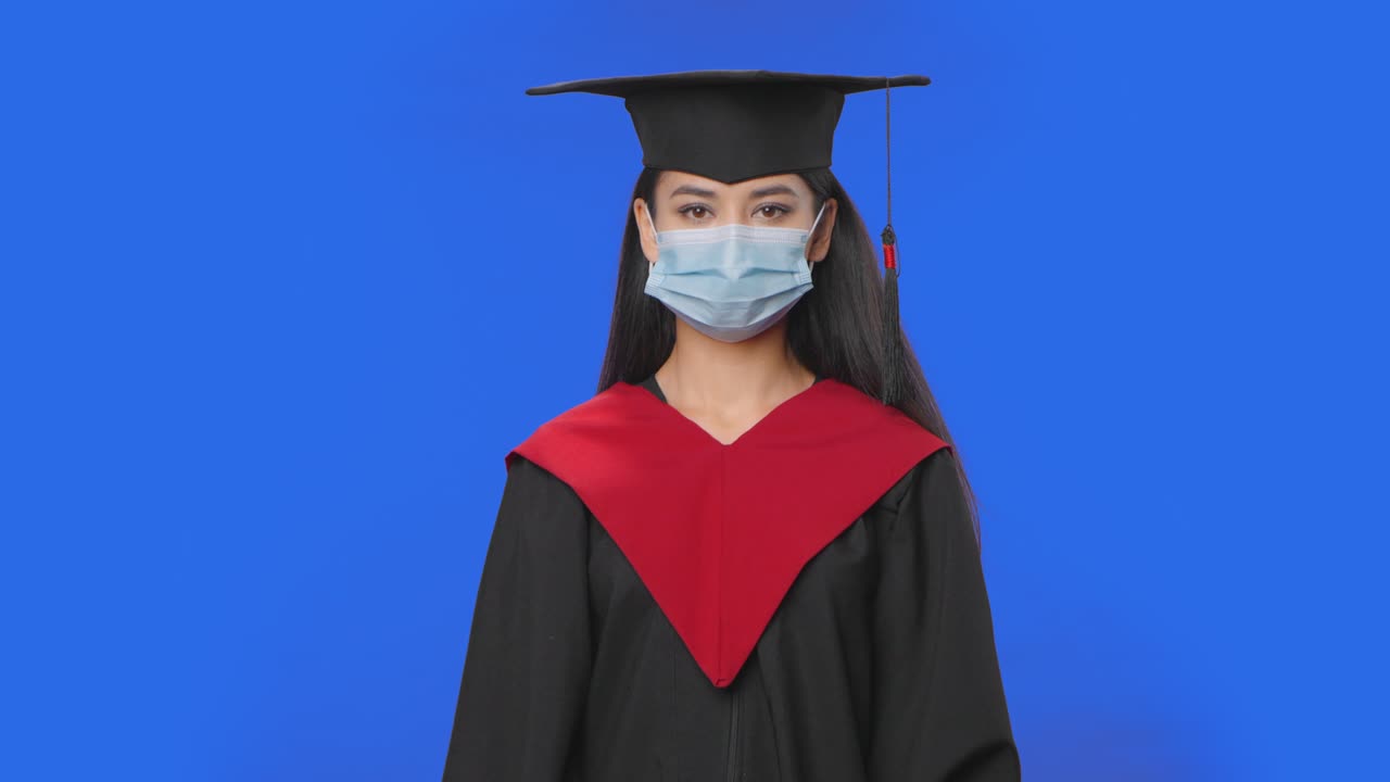 Portrait of female student in cap, gown graduation costume and medical mask looking at camera. Young brunette woman posing in studio with blue screen background. Close up. Slow motion ready 59.94fps
