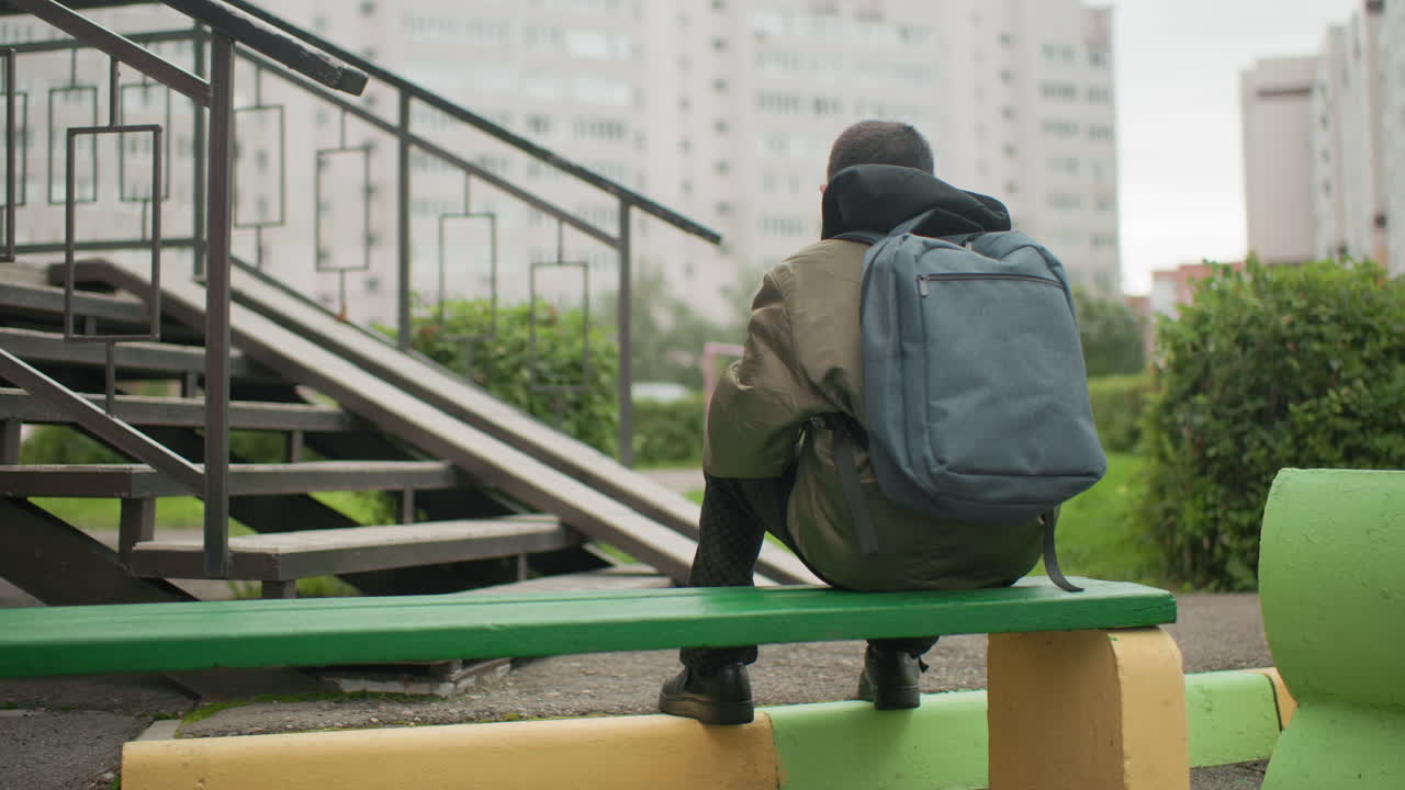 Rear view of boy seated on green bench wearing backpack while studying outdoors near residential buildings with blurred background and stair railing showing focus