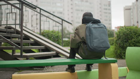 rear view of boy seated on green bench wearing backpack while studying outdoors near residential buildings with blurred background and stair railing showing focus