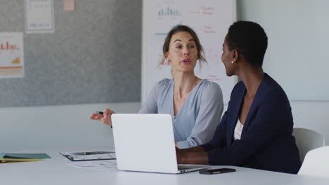 dos colegas femeninas diversas sentadas en el escritorio con una computadora portátil y discutiendo en la oficina