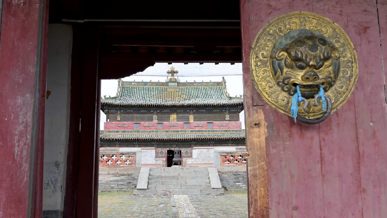 Looking through a historic gateway at Erdene Zuu. The main Buddhist temple with its green tiled roof is revealed behind a traditional brass door handle and ceremonial scarf