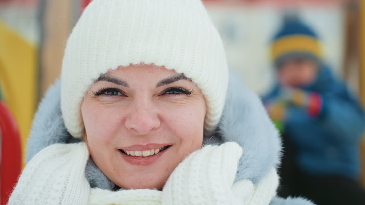 Mamma e figlio che si lanciano palle di neve in un parco giochi, sequenza di reazioni spontanee, donna con berretto di lana bianco e sciarpa che reagisce dalla sorpresa al sorriso, bambino sfocato che lancia neve in un colorato parco giochi all'aperto, dinamico.