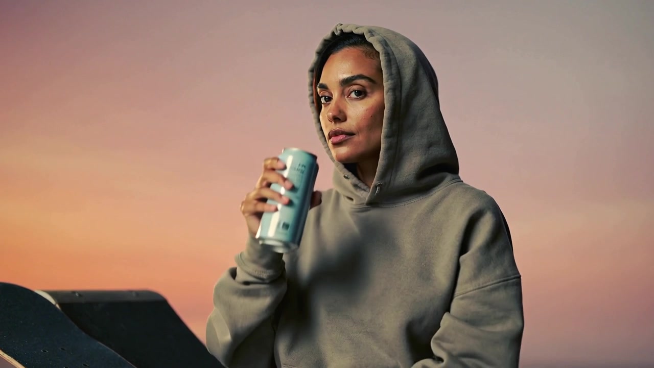 Una donna che apre la linguetta di una lattina, con felpa grigia con cappuccio in studio, che sorseggia da una lattina blu per idratarsi, skateboard a sinistra