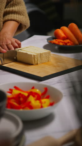 vertical video close up of woman at home in kitchen preparing healthy vegetarian or vegan meal slicing tofu on board with knife 3