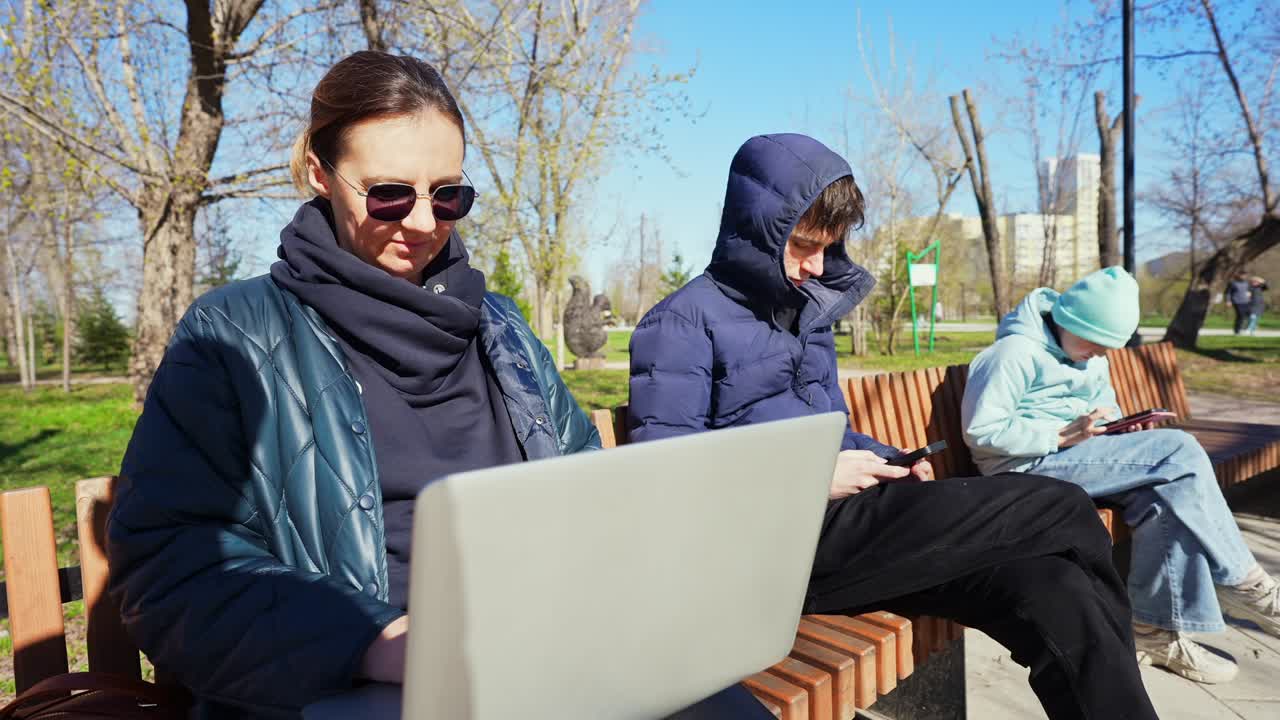 Kvinde arbejder på laptop med børn, der bruger enheder på en parkbænk