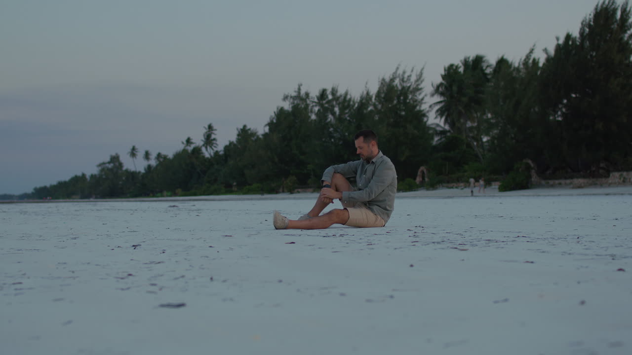 Parallax shot of Lonely man sits in silence on pure white sand tropical beach contemplating the beauty of the ocean during twilight.