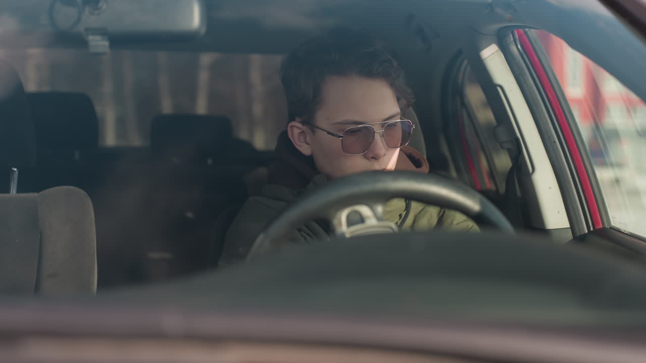 young man inside parked car seen through windshield removing seatbelt while holding steering wheel and looking downward with focus as winter sunlight streams through windshield