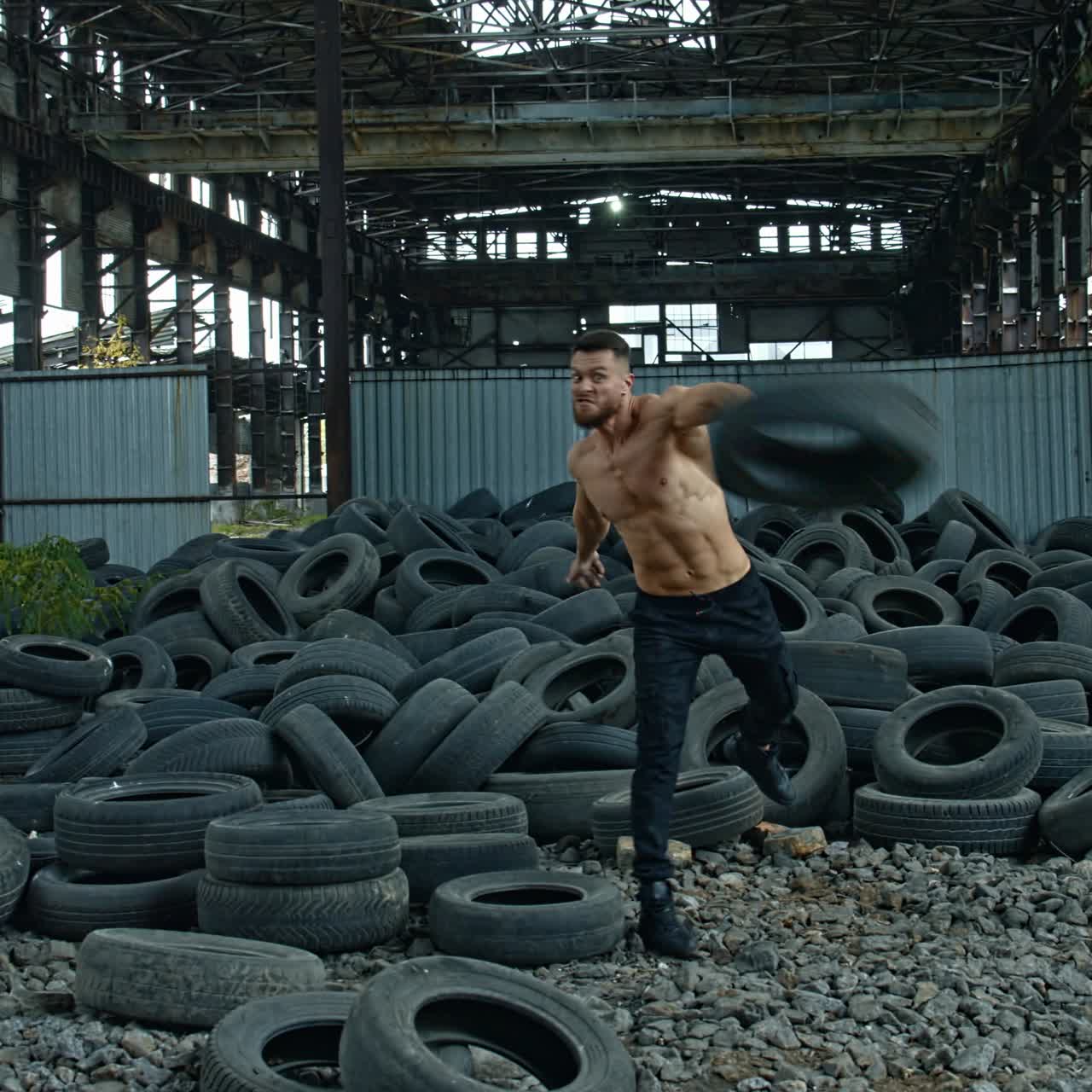 Training session of a sportsman in abandoned place. Strong man without shirt scattering over old car tires during his workout on ruined factory background.