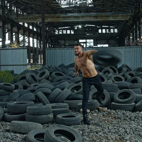 training session of a sportsman in abandoned place. strong man without shirt scattering over old car tires during his workout on ruined factory background.
