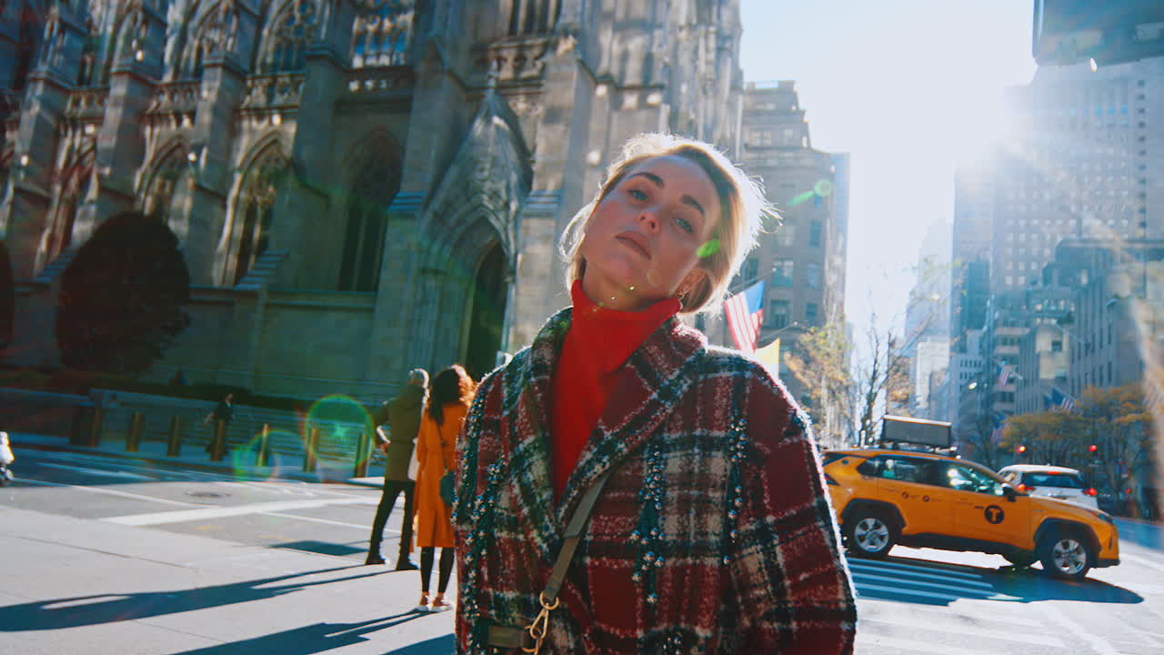 Nueva York, EE. UU., 2 de abril de 2026: Mujer a la moda posando cerca de la Catedral de San Patricio en Nueva York. Mujer con abrigo de cuadros rojo y guantes verdes sosteniendo gafas de sol con catedral gótica y destello de lente