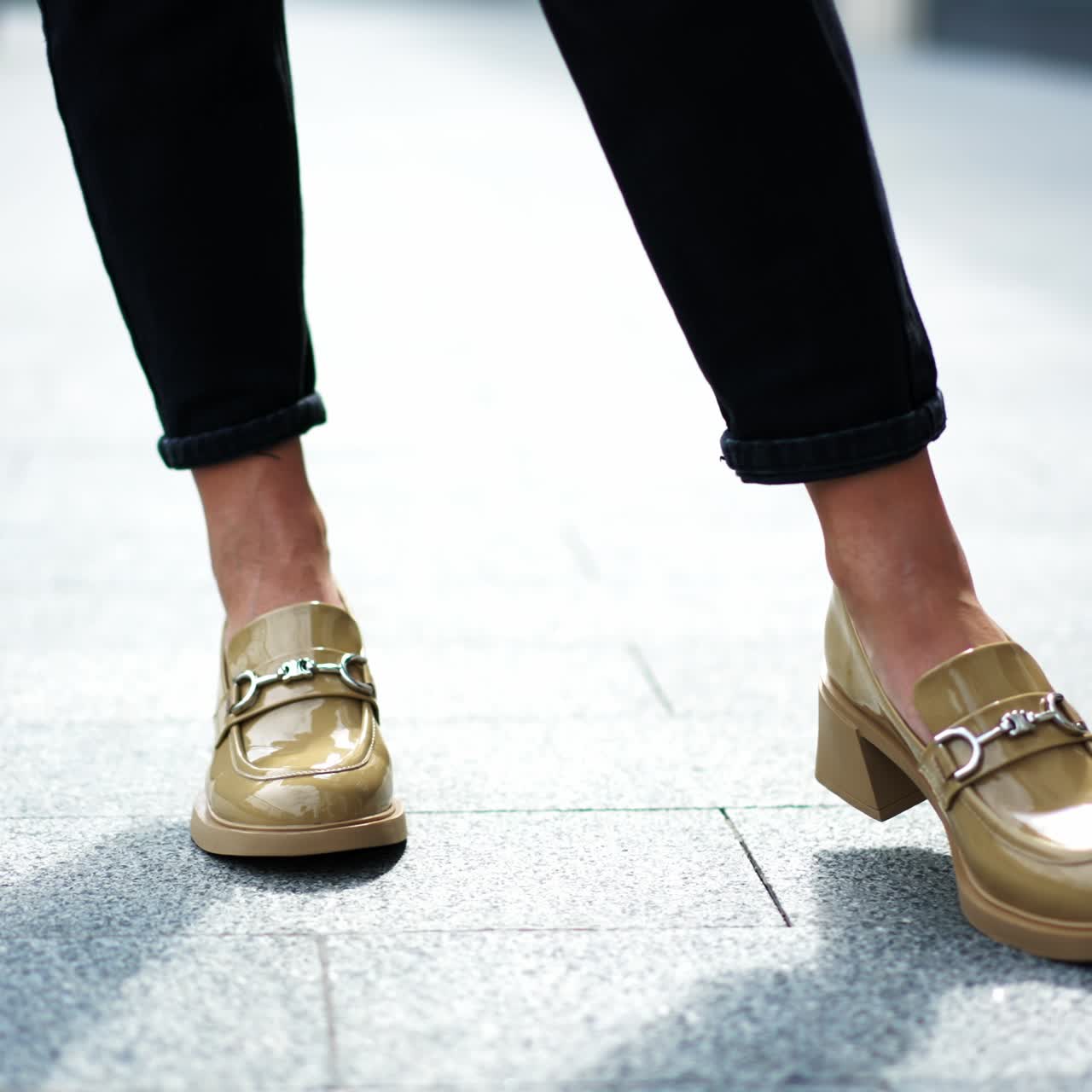 Beige patent leather shoes with metal decoration. Female model in black pants demonstrating the footwear. Close up