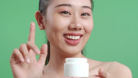 미소 짓는 아시아 여성이 스튜디오에서 녹색 화면 배경에 서서 카메라에 얼굴 크림을 보여주고 있습니다.