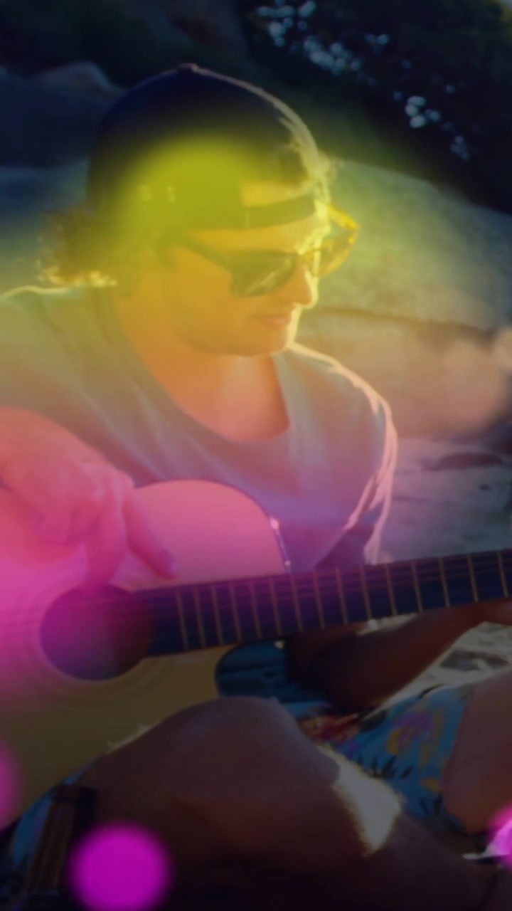 Vídeo vertical: Guitarrista dedilhando na praia enquanto um amigo passa uma garrafa, com bokeh rosa sobre a cena para música.