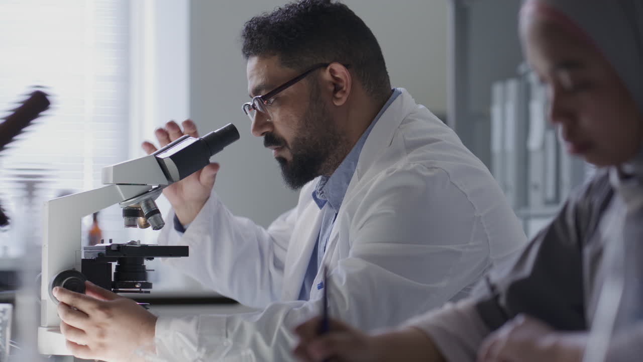 Scienziato al microscopio che prende appunti accanto a una collega in laboratorio