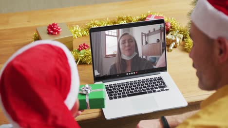 Padre-E-Hijo-Caucásicos-Con-Gorros-De-Papá-Noel-Usando-Una-Computadora-Portátil-Para-Una-Videollamada-Navideña-Con-Una-Mujer-En-La-Pantalla