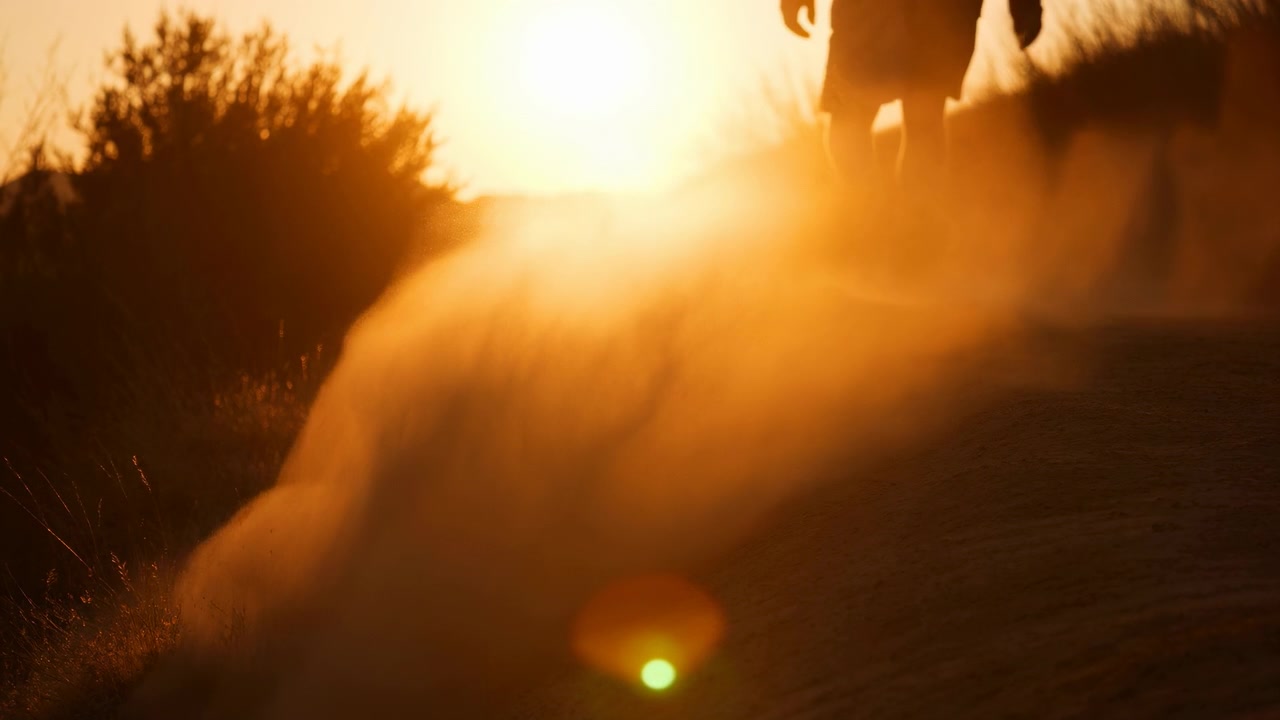 Parte inferior das pernas em shorts caminha por uma trilha arenosa enquanto o impacto do pé levanta poeira em uma pluma aérea brilhante, forte contraluz do pôr do sol cria uma luz de contorno âmbar quente, sombra suave e aventura ao ar livre