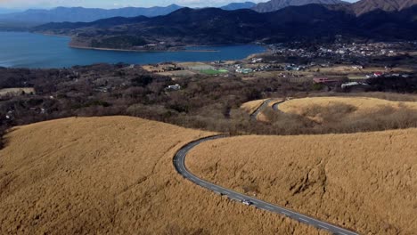 A-winding-road-through-golden-fields-with-a-lake-and-mountains-in-the-distance,-under-clear-skies,-aerial-view