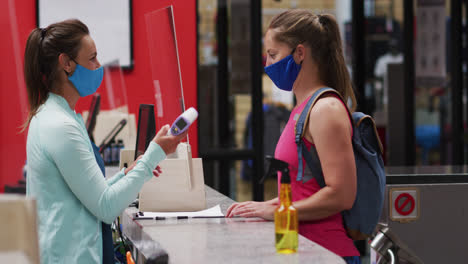 caucasian female gym receptionist in face mask taking temperature of masked female client