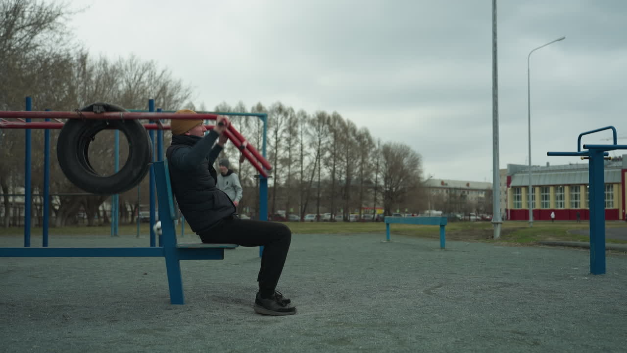 A coach is seen taking a break from his workout on outdoor equipment in a stadium, in the background, two boys play football, while buildings, parked cars seen at a distance
