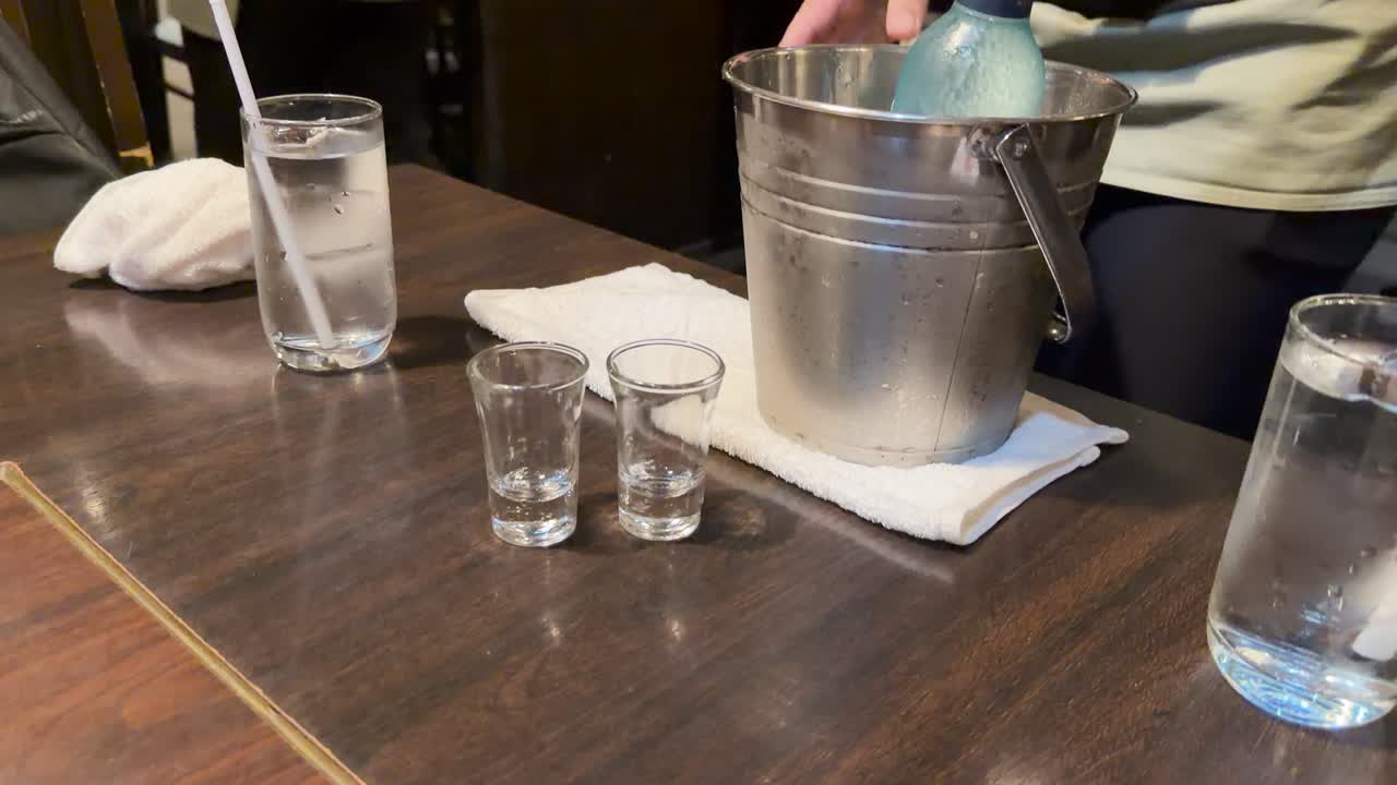 Server places shot glasses next to ice bucket with sake bottle on wooden restaurant table