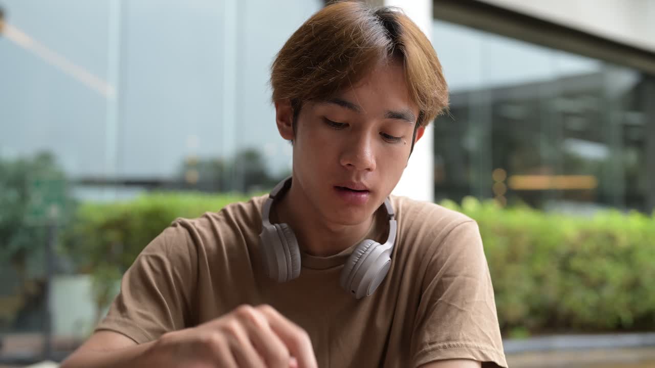 Young handsome Asian man sitting and eating french fries while sitting in outdoors restaurant during summer