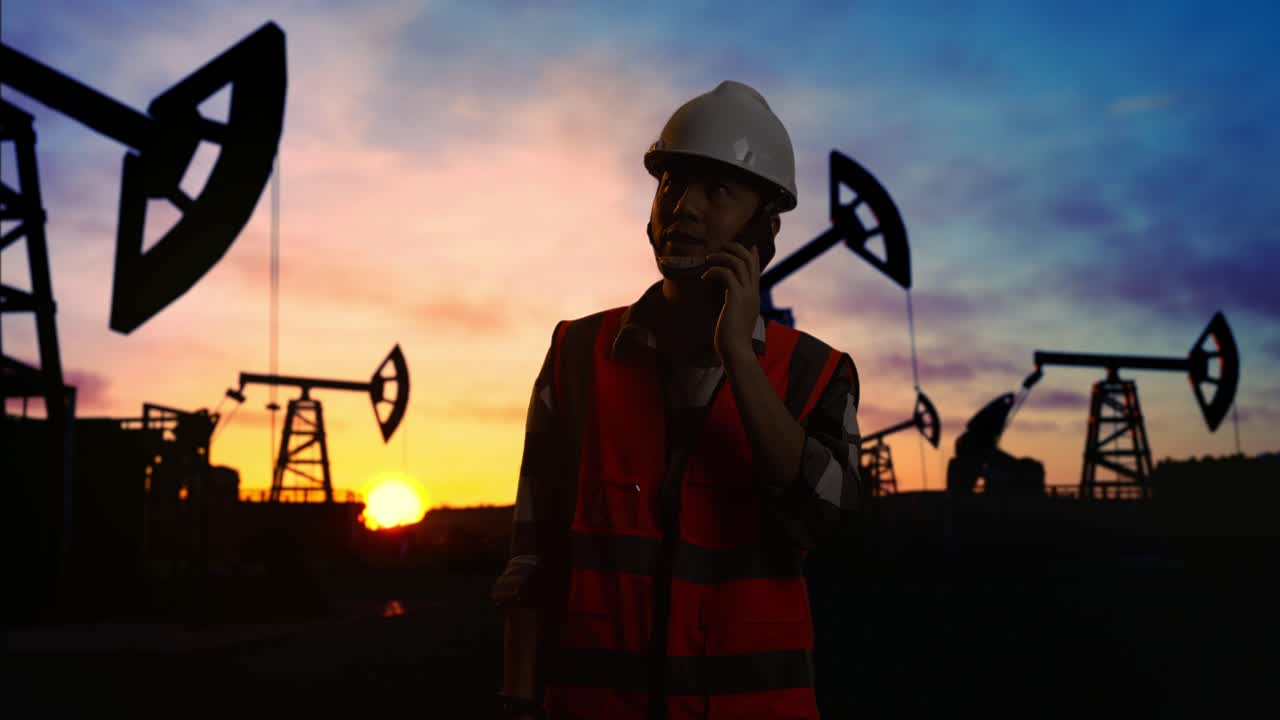 ingeniero masculino asiático con casco de seguridad hablando en teléfono inteligente mientras está de pie frente a las bombas de aceite, durante la puesta o el amanecer
