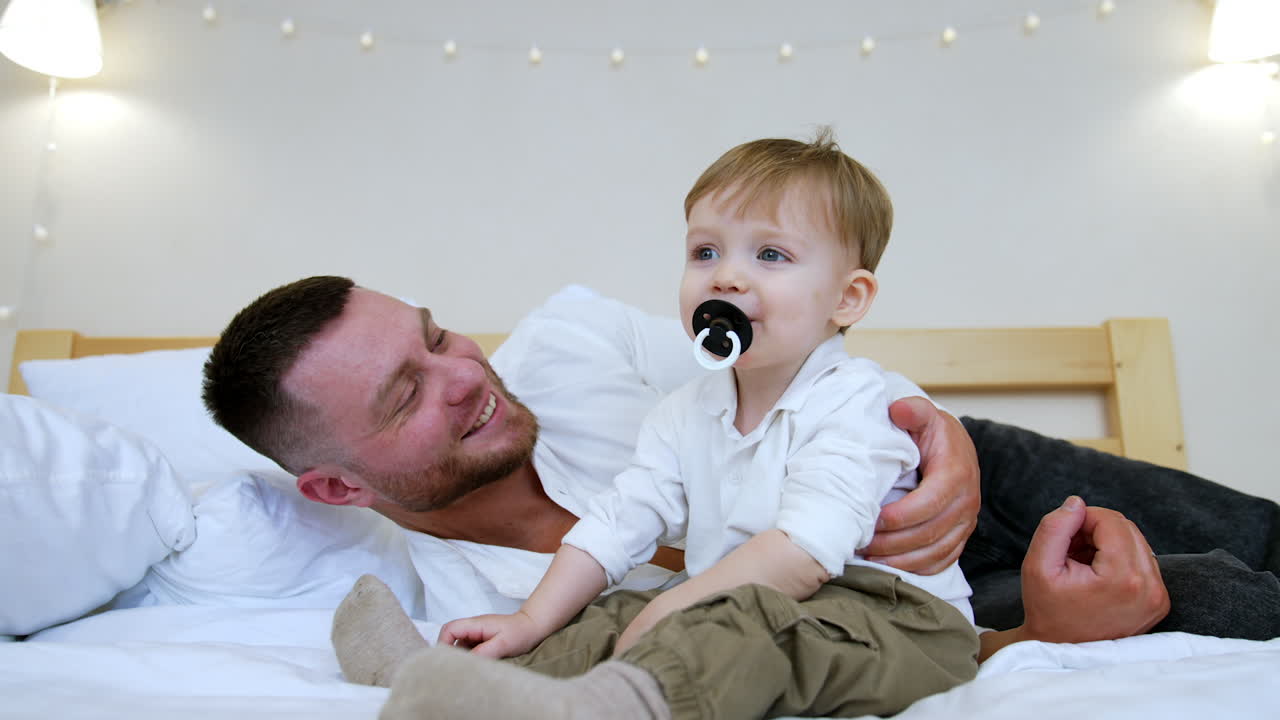 Beautiful baby boy with pacifier in mouth sits on the bed near his dad. Loving father kisses his adorable son on the cheek. Man wants to take away a pacifier but the kid protests and gets it again.