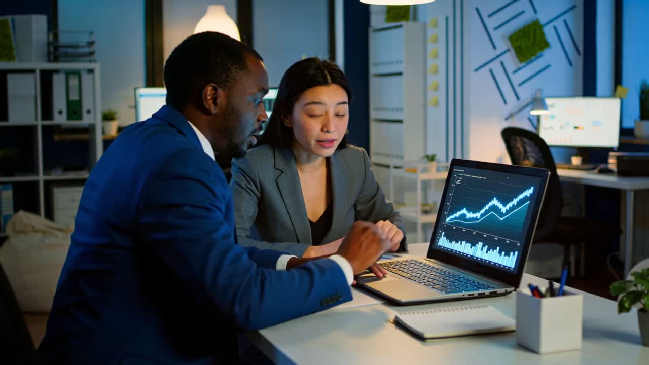 Noticing upward trend on laptop at desk pointing pair in suits examining chart and taking notes