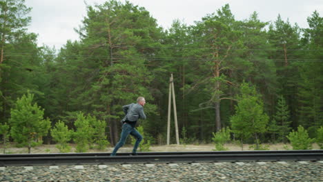 a man in a grey suit jacket and blue jeans is seen running and looking back energetically along railway tracks, surrounded by lush green forest and yellow poles