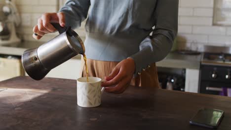 Midsection-of-caucasian-woman-standing-at-counter-in-cottage-kitchen-pouring-coffee-from-pot