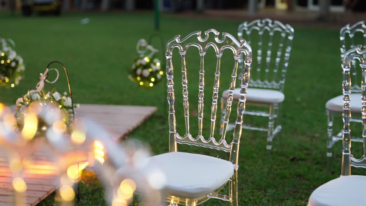 Lenta panoramica su sedie trasparenti in acrilico su un prato verde accanto a un corridoio nuziale decorato, con luci bokeh soffuse e accenti floreali, che rivelano un'elegante disposizione dei posti a sedere e un'atmosfera esterna raffinata