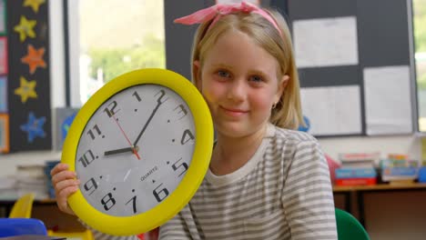 Vorderansicht-Eines-Kaukasischen-Schulmädchens-Mit-Wanduhr,-Das-Am-Schreibtisch-Im-Klassenzimmer-Sitzt-4k