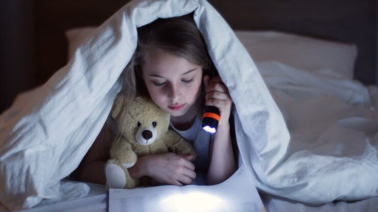 Cute little girl hugging a teddy bear, using a flashlight and reading a book with surprised expression under the blanket at night
